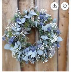 a wreath with blue flowers and greenery hanging on a wooden fence next to a rope