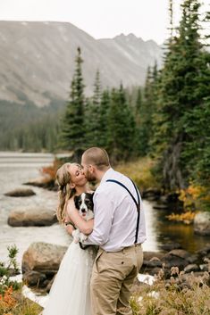 a bride and groom kissing in front of a mountain lake with their dog on their wedding day