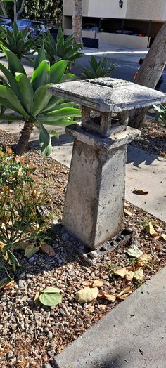 there is a small stone statue in the middle of some rocks and plants on the side walk