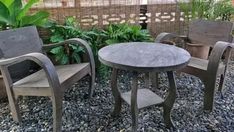 three chairs and a table sitting on gravel in front of a fenced area with plants