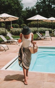 a woman in a hat is walking near a pool with chairs and umbrellas around her