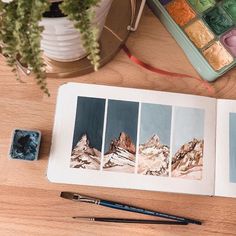 an open book sitting on top of a wooden table next to watercolor paints and a brush