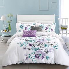 a white bed with purple flowers on the comforter and pillows in front of a blue wall