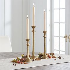 three candles are sitting on a table with leaves and berries around it, in front of a window