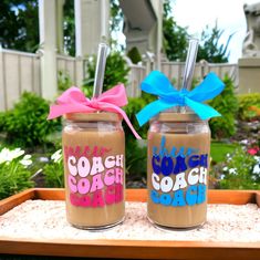 two glasses with straws on top of sand in front of some flowers and trees