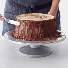 a person holding a knife in front of a cake on a platter with chocolate frosting