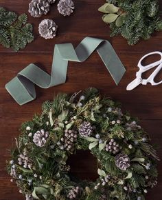 the wreath is being made with green tape and evergreen leaves, pine cones, berries, and scissors