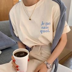 a woman sitting on a couch holding a cup of coffee and wearing a white t - shirt that says je me sais quijo