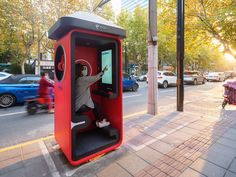 a man standing in a red phone booth on the side of a street with cars