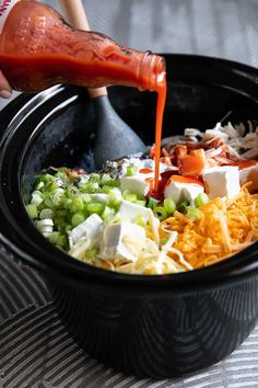 someone pouring dressing into a black bowl filled with vegetables and cheese, including celery