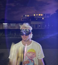 a young man with blonde hair and goggles standing in front of a building at night