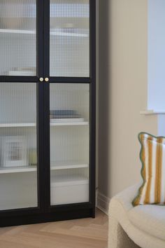 a black cabinet with glass doors in the corner next to a white chair and pillow