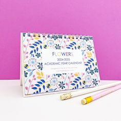 a calendar and two pencils sitting on a table next to a pink background with flowers