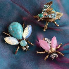 three insect brooches sitting on top of a blue cloth covered in beads and stones