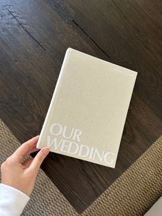 a person holding up a wedding book on top of a wooden floor with the words our wedding printed on it