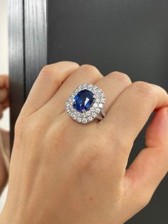 a woman's hand holding an oval shaped blue and white diamond ring in front of a mirror