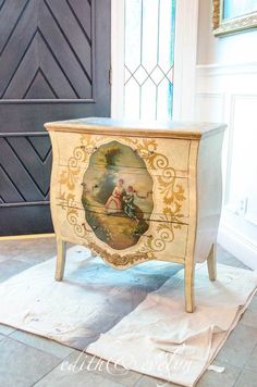 an old dresser with a painting on the front and side drawers painted white, sitting next to a black door