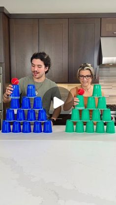 two people standing in front of a kitchen counter with blue and green cups stacked on top of each other