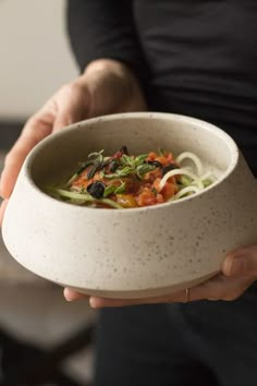 a person holding a bowl filled with noodles and veggies on top of it