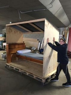 a man standing next to a wooden bed in a warehouse with mattresses on it