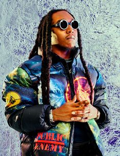 a man with dreadlocks standing in front of a wall wearing a jacket and sunglasses