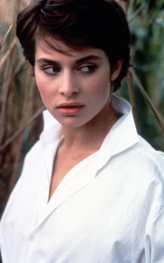 a woman with short hair wearing a white shirt and looking at the camera while standing in front of trees