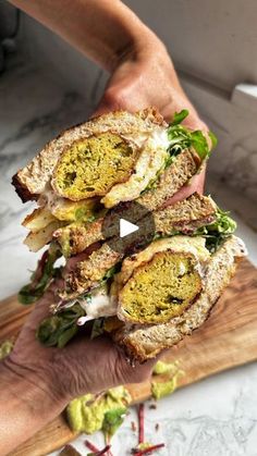 a person is holding a sandwich on a cutting board with other food items in the background