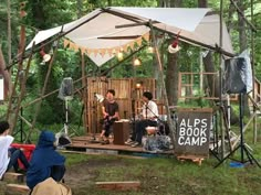 some people are sitting in the woods and playing music under a tent with lights hanging from it