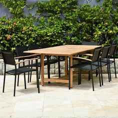 a wooden table with black chairs in front of some bushes and plants on the side walk