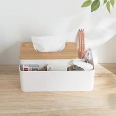 a white box with some pens and pencils in it on a wooden table next to a potted plant