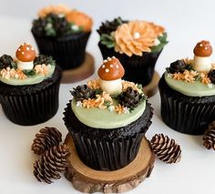 four cupcakes decorated with mushrooms and flowers on top of a wooden stand next to pine cones