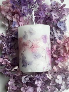 a white candle with purple flowers on it sitting next to some pink and purple leaves