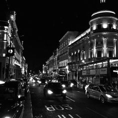 black and white photograph of cars driving down the street at night time with buildings in the background