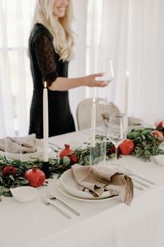 a woman standing next to a table set with plates and silverware, candles and greenery