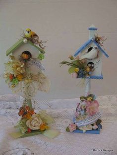 two small birdhouses with flowers and birds on them sitting on a lace tablecloth