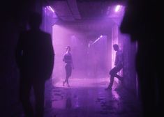 three people walking in a dark tunnel with purple light coming from the ceiling and floor