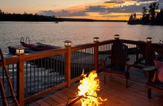 a fire pit sitting on top of a wooden deck next to a lake at sunset