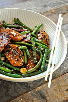 a white bowl filled with green beans and meat covered in sesame seeds next to chopsticks