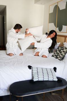 two people in white robes are on a bed and one person is holding up pillows