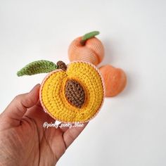 a hand holding an orange crocheted brooch with two pieces of fruit in the background