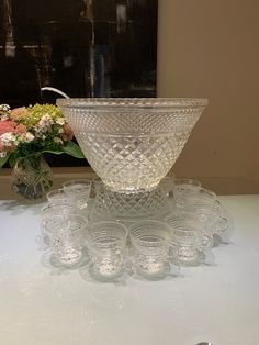 a clear glass bowl and some glasses on a white table with flowers in the background