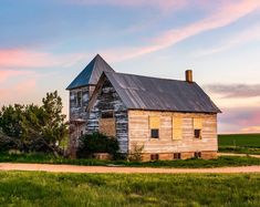 an old run down house sitting in the middle of a green field with a sunset behind it