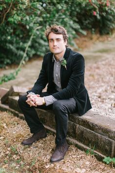 a man in a suit and tie sitting on a stone wall with trees behind him