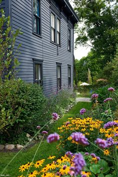 a house that is next to some flowers in the grass and bushes on the side