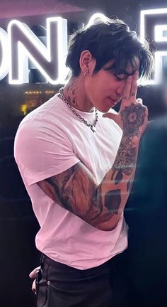 a man with tattoos standing in front of a neon sign holding his hand to his face