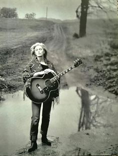 a woman holding a guitar standing next to a body of water in the grass and trees