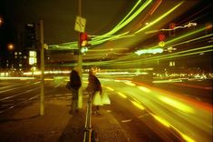 two people are standing on the side walk at night with traffic lights in the background