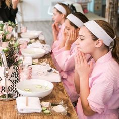 women in pink robes sitting at a long table with plates and bowls on it, all covering their faces