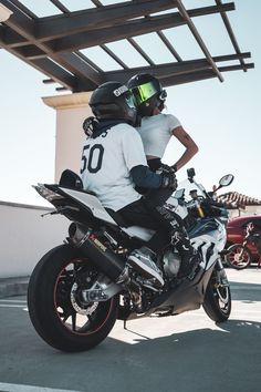 two people sitting on the back of a motorcycle in front of a building with a sky background