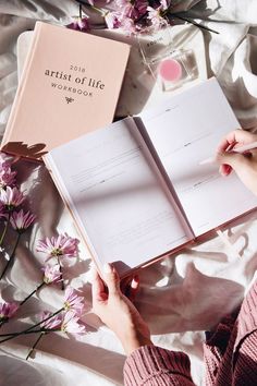 a woman is holding an open book and writing on the pages next to some flowers